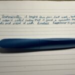 A photo of an aluminum blue Pelikan Twist Fountain Pen against white paper with handwriting. The handwriting reads: “Technically, I bought this pen last week, but it wasn’t until today that I found a converter that fit and inked it with Kuretake Kujakuryoku.