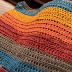 A crocheted blanket made by using the half-double crochet stitch with multi-colored yarn displaying the following colors from the bottom up: teal, gray, red, orange, yellow, and teal.